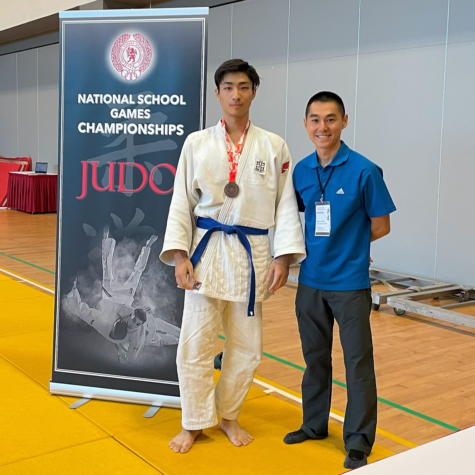 A judoka in a white gi with a medal stands with an official beside a National School Games Championships Judo banner.